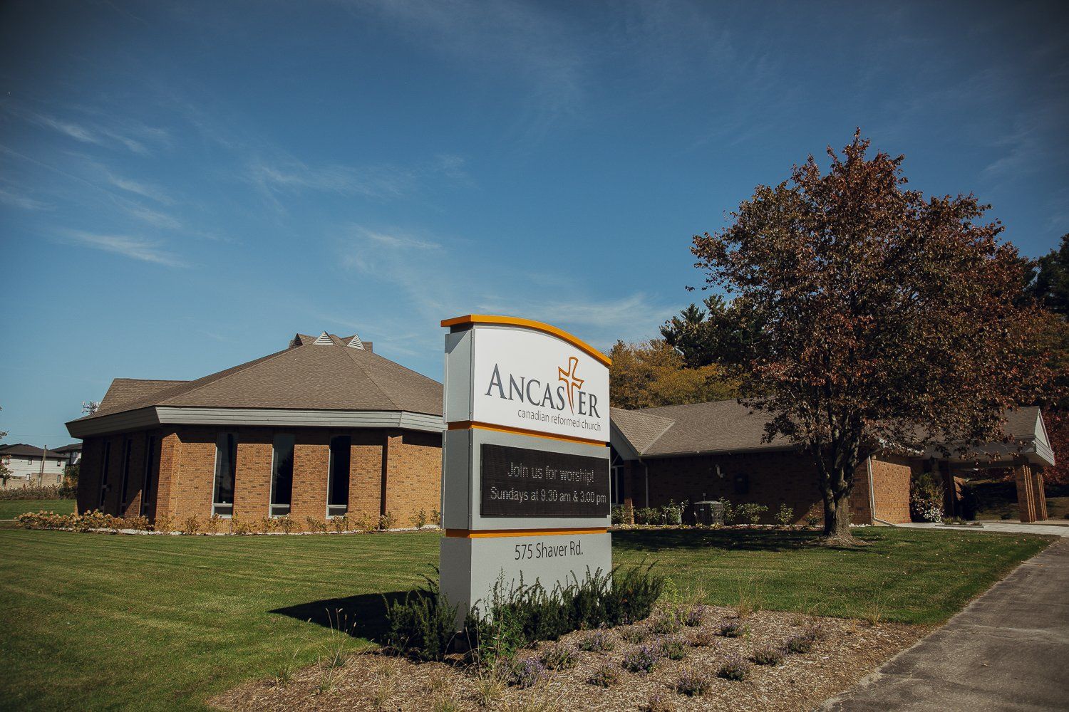 Ancaster Canadian Reformed Church, a long, low brick building with a brown hipped roof beyond a lawn and a tree with dark red leaves, behind the church's sign giving its Sunday service times and its address, 575 Shaver Road