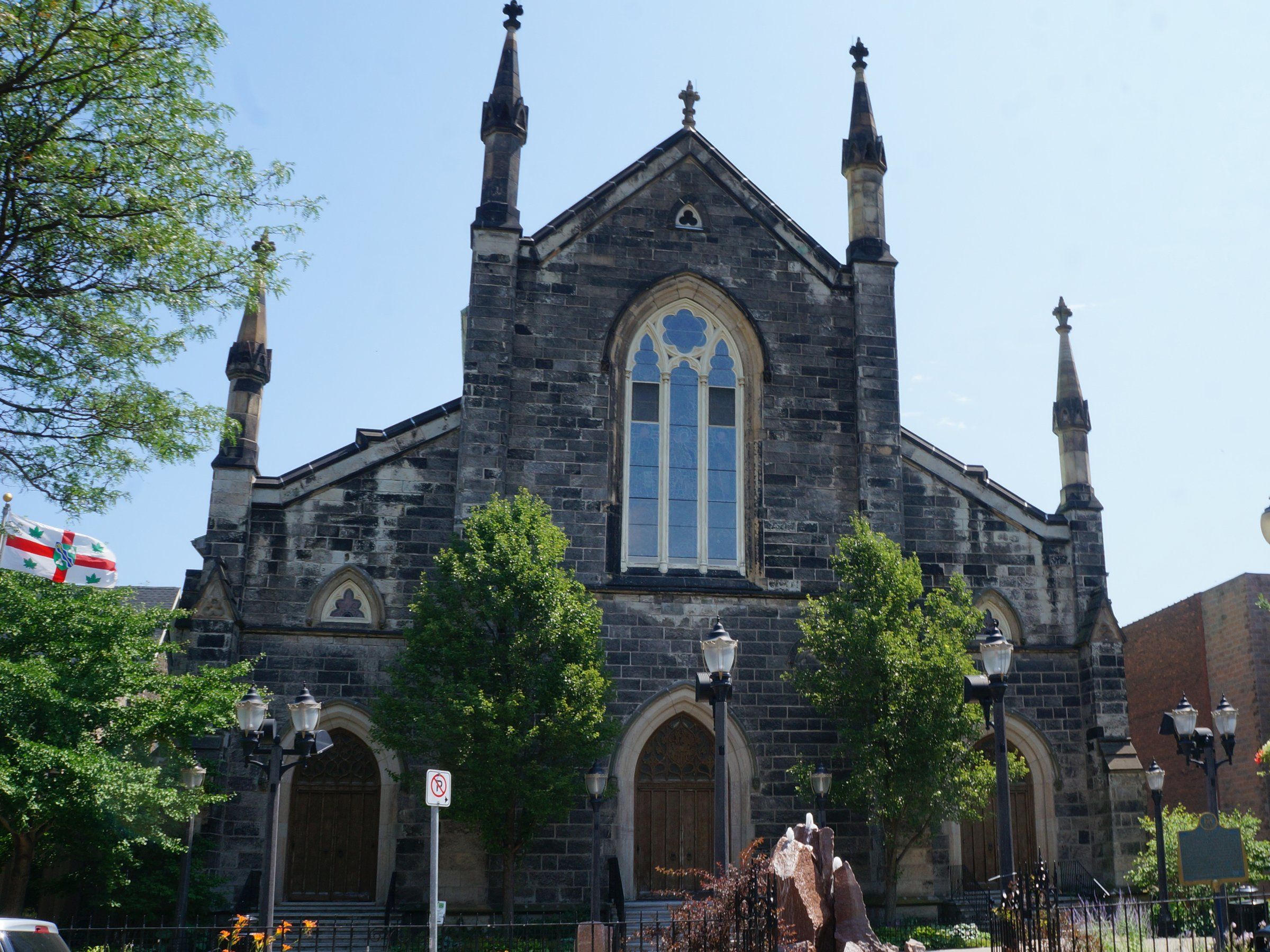 Christ's Church Cathedral, a dark grey stone church with a tall pointed window above pointed-arch doorways and slender pinnacles at its corners, seen from James Street North behind young trees