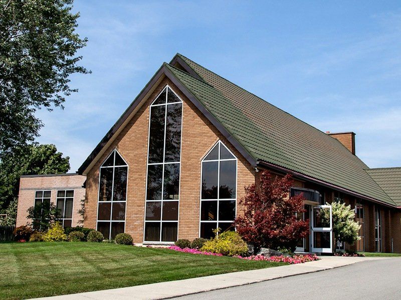Cornerstone Canadian Reformed Church, a tan-brick church with a steep green roof and three tall pointed windows in its gable end, behind a lawn and flower beds