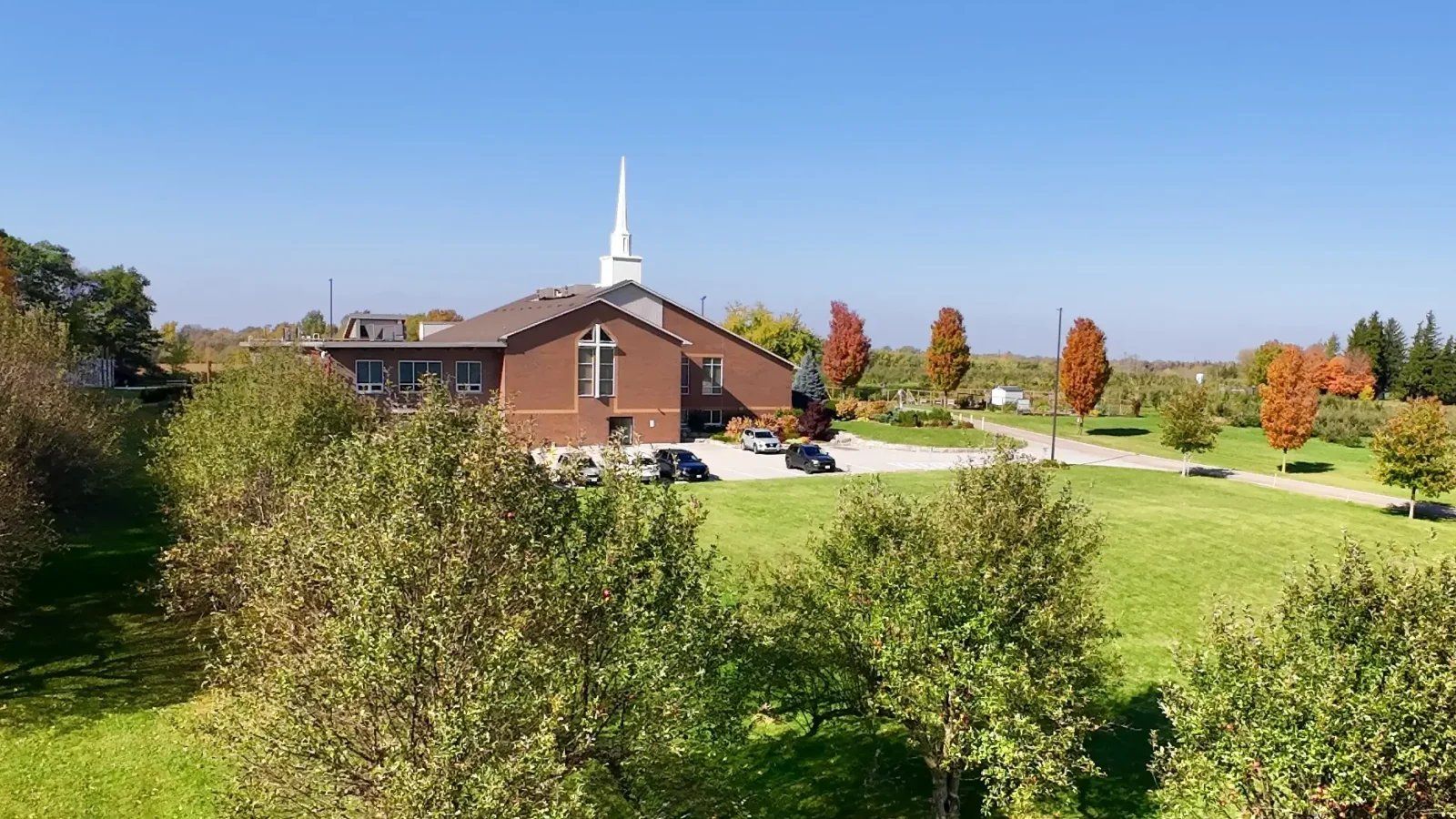 Flamborough Baptist Church, a red-brick church with a tall white steeple and a large window divided by a cross in its front gable, seen across a wide lawn and trees in autumn colour