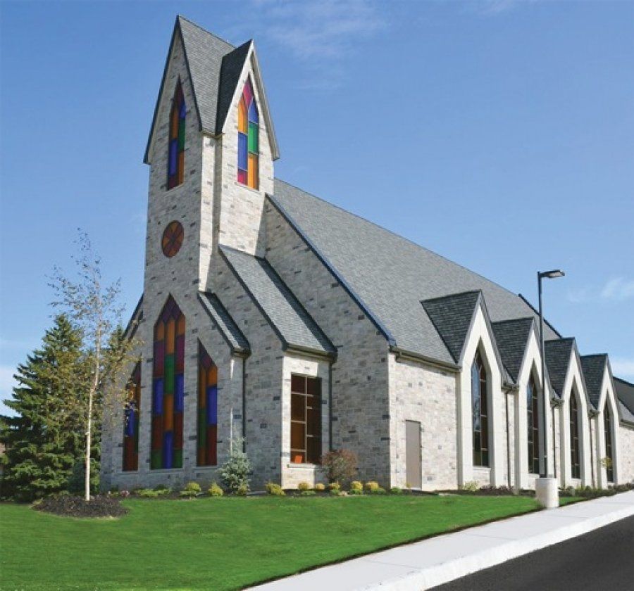 Maranatha Free Reformed Church, a pale grey church with a tall gabled tower set with stained glass, tall stained-glass windows and a row of pointed gables along its side, above a lawn and a sidewalk