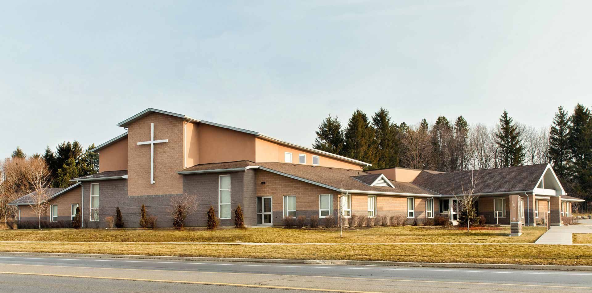 The Salvation Army Meadowlands Community Church, a long single-storey building in tan and grey-brown block with a taller gabled sanctuary bearing a large white cross and a peaked entrance canopy, set back behind a wide lawn