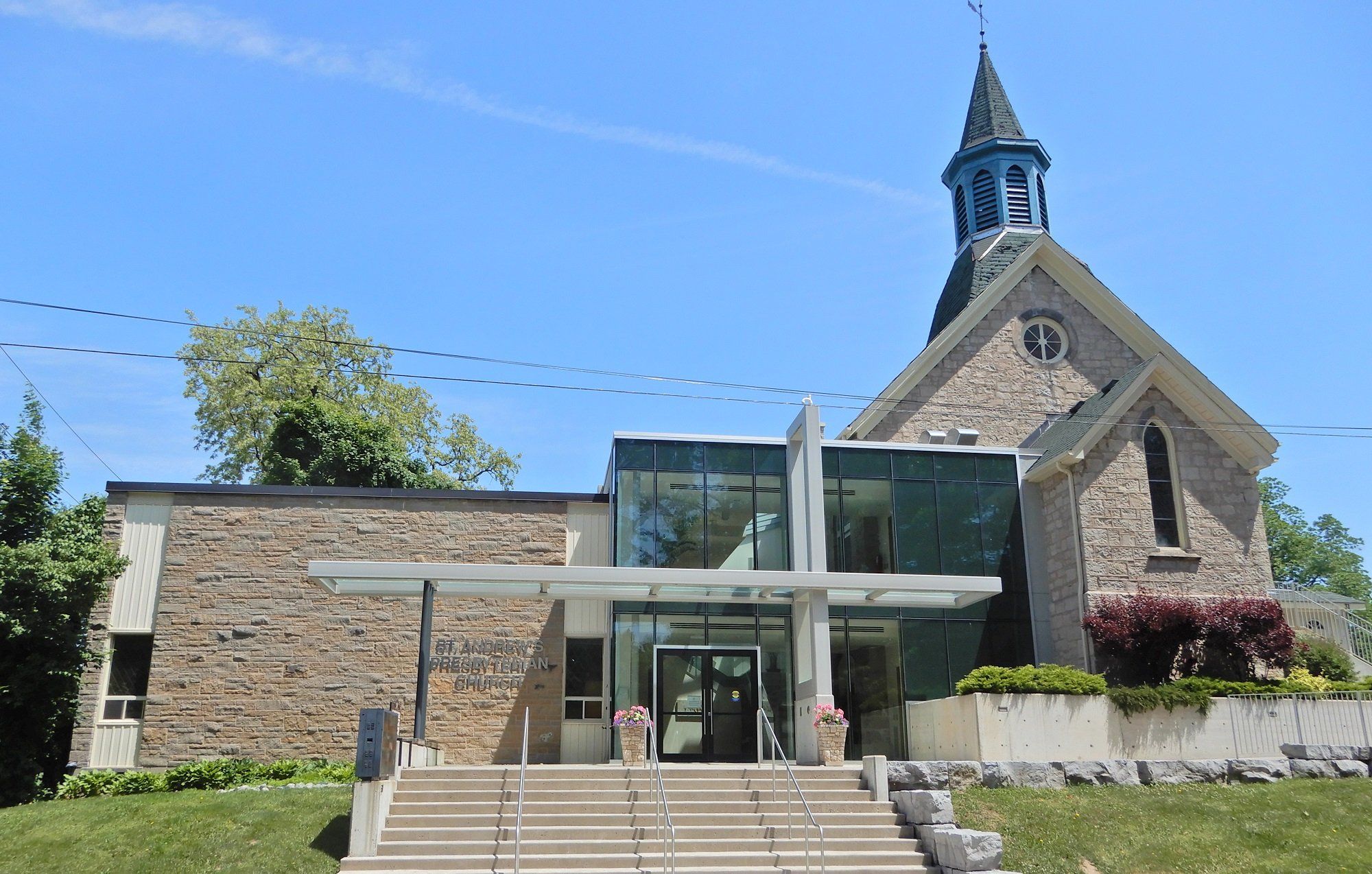 St. Andrew's Presbyterian Church, a grey stone church with a round window and a small bell tower with a dark spire, joined by a glass entrance lobby to a lower stone wing lettered with the church's name, above a wide flight of steps