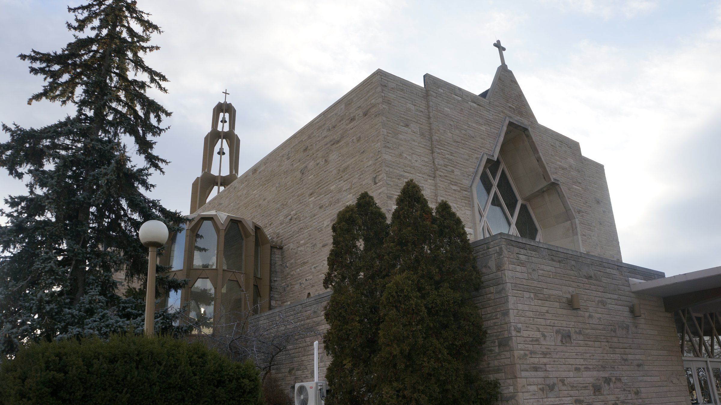 St. Francis Xavier Church, a pale stone church with a tall pointed window, a round glass-walled bay and an open bell tower, behind evergreens