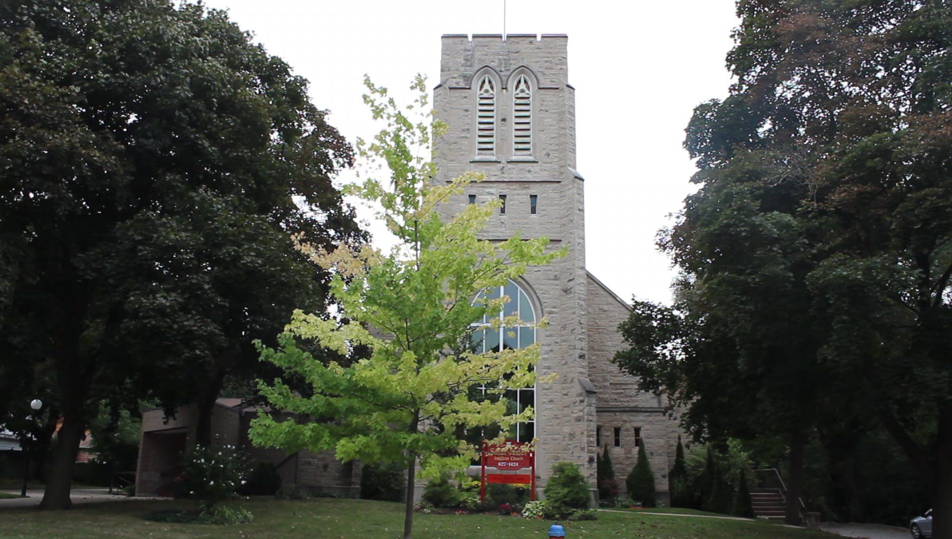 St. James Anglican Church, a grey stone church with a tall square tower, pointed belfry openings and a large arched window, behind a young tree and a red parish sign