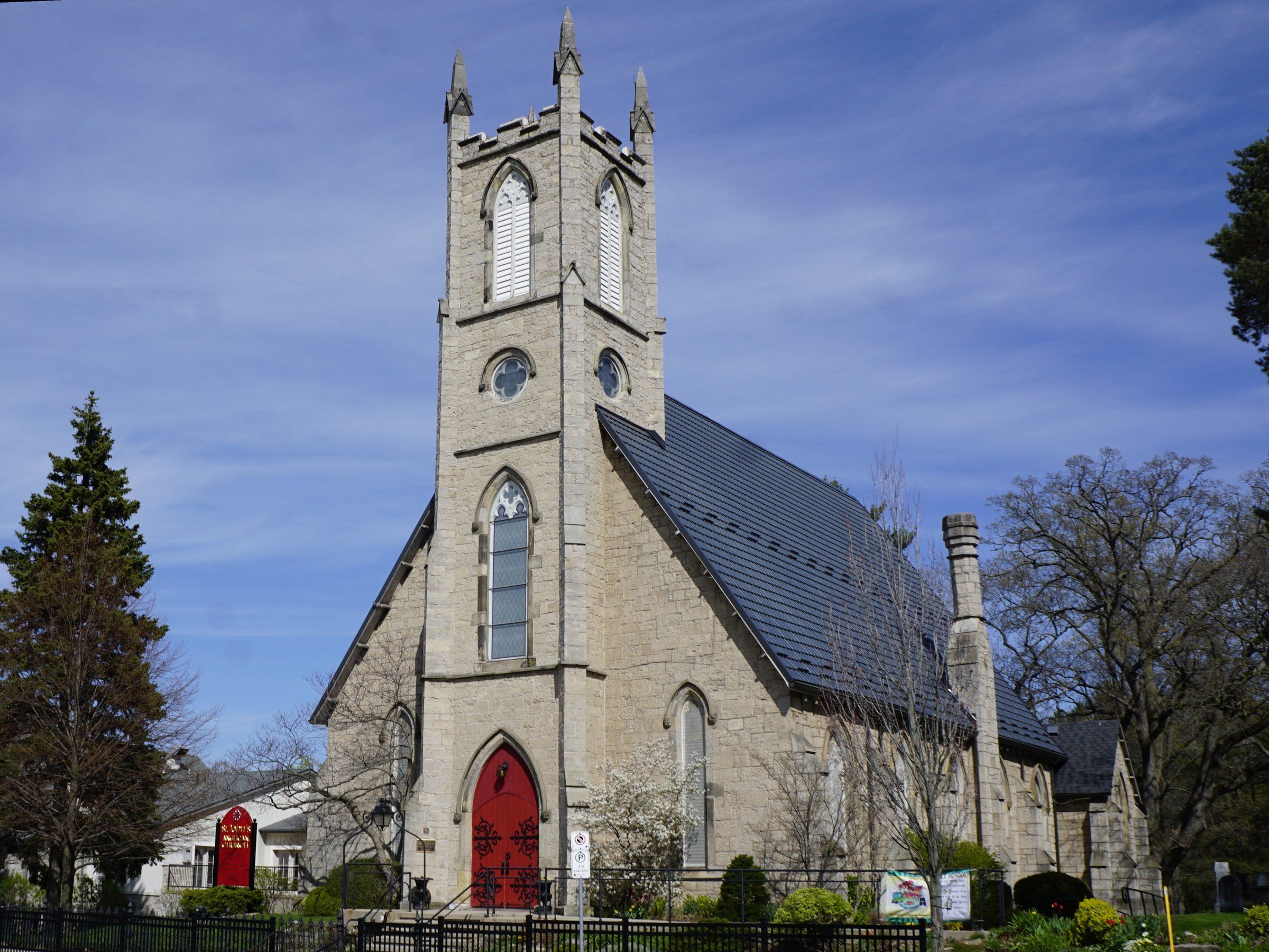St. John's Anglican Church, a grey stone church with a square pinnacled tower above a red pointed-arch door and a steep dark roof, behind an iron fence