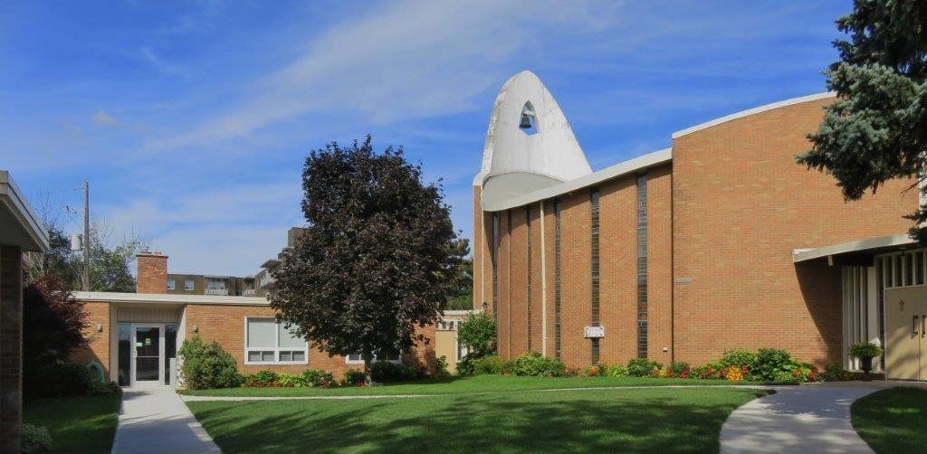 St. Michael's Anglican Church, a curved buff-brick church with tall narrow windows and a white rounded concrete bell tower, beside a low brick hall and a lawn crossed by paths