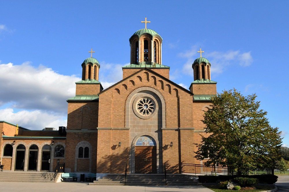 St. Nicholas Serbian Orthodox Cathedral, an orange-brown brick church with three green domes topped by gold crosses, a round window and an icon above its doors, and a tree at its side