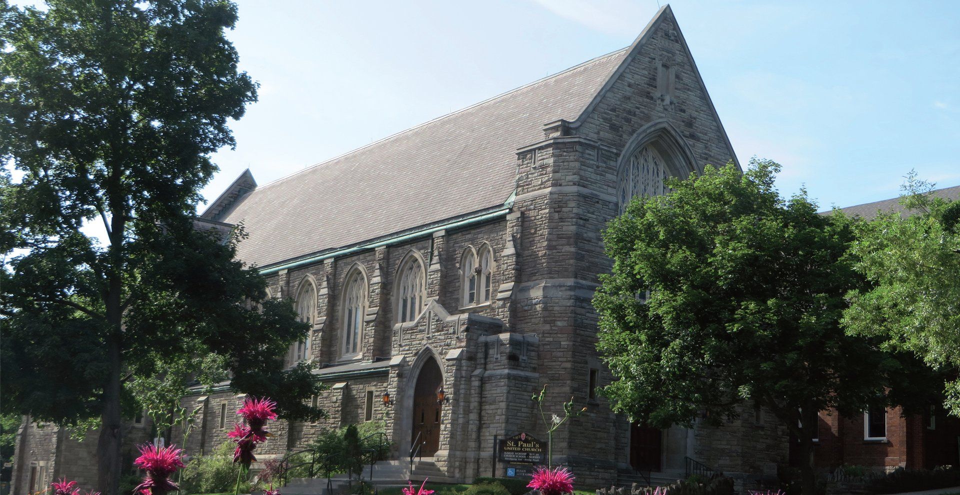 St. Paul's United Church, a grey stone church with a steep grey roof, a large traceried window in its gable and a pointed-arch door, behind trees and pink flowers, with its sign on the lawn