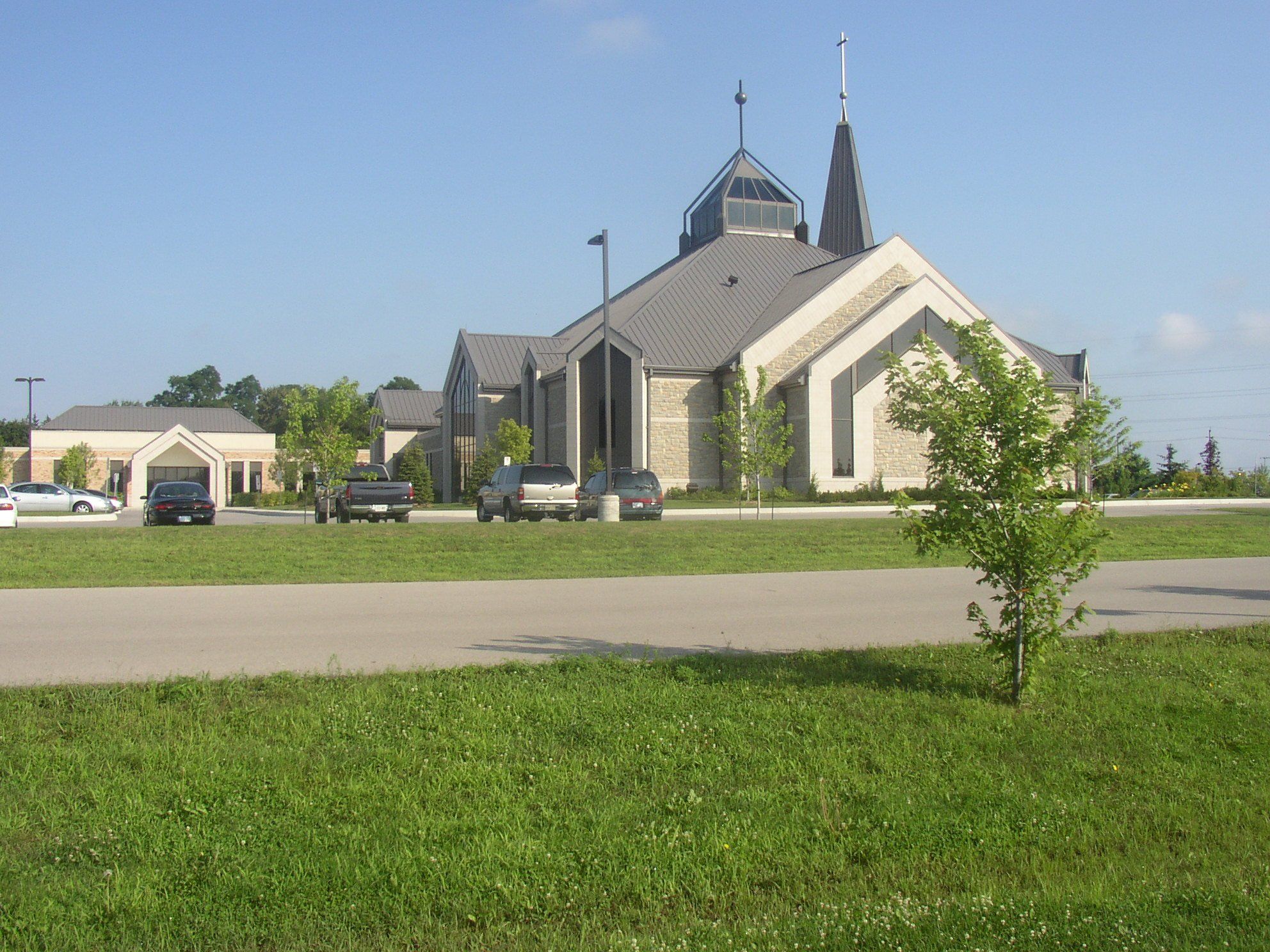 St. Thomas the Apostle Church, a large pale stone church with steep grey roofs, a glazed lantern and a slender dark spire, seen across a lawn and its parking lot