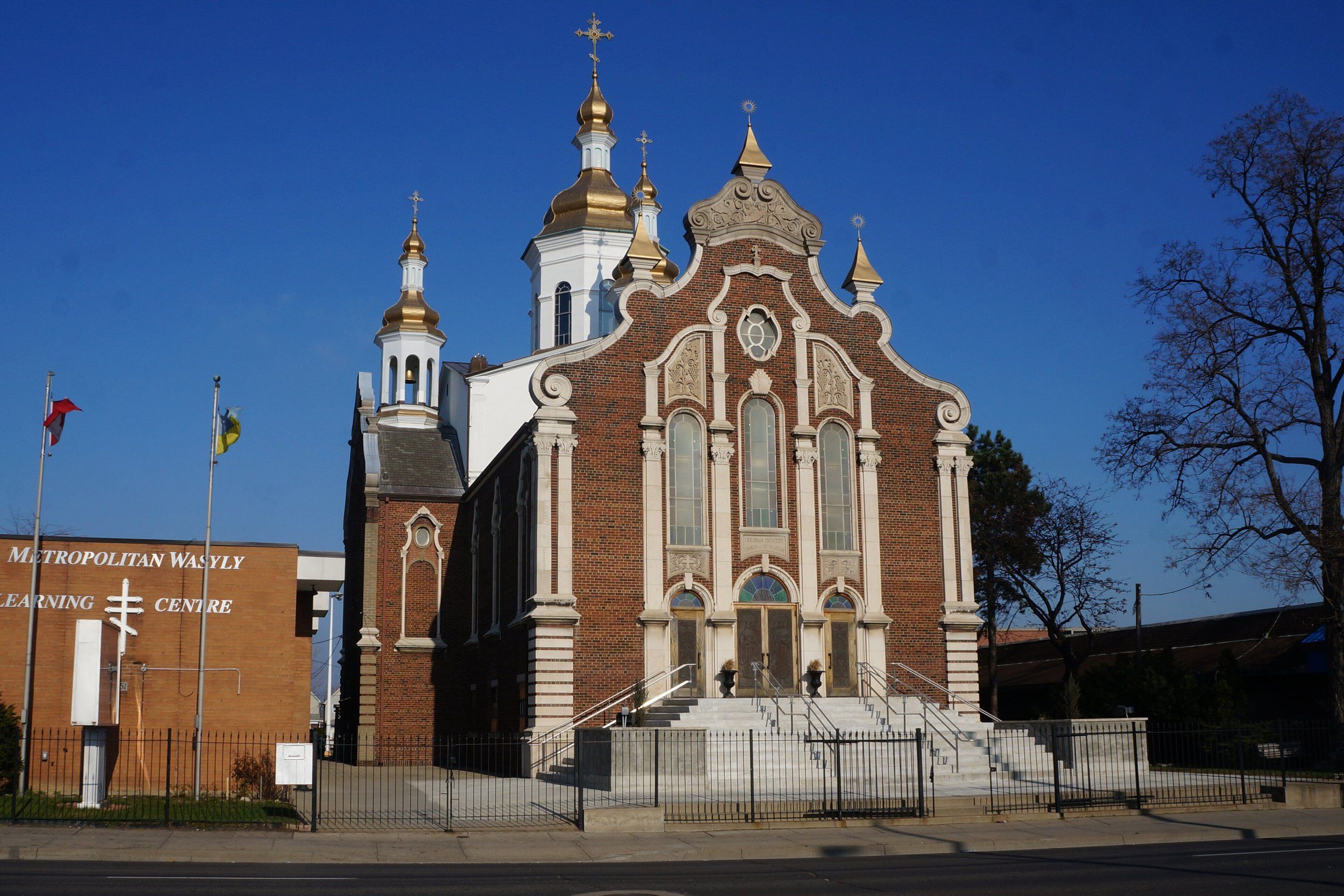 The Ukrainian Orthodox Cathedral of St. Vladimir, a red-brick church with a pale stone-trimmed front, tall arched windows and gold onion domes topped with crosses, behind an iron fence on Barton Street East