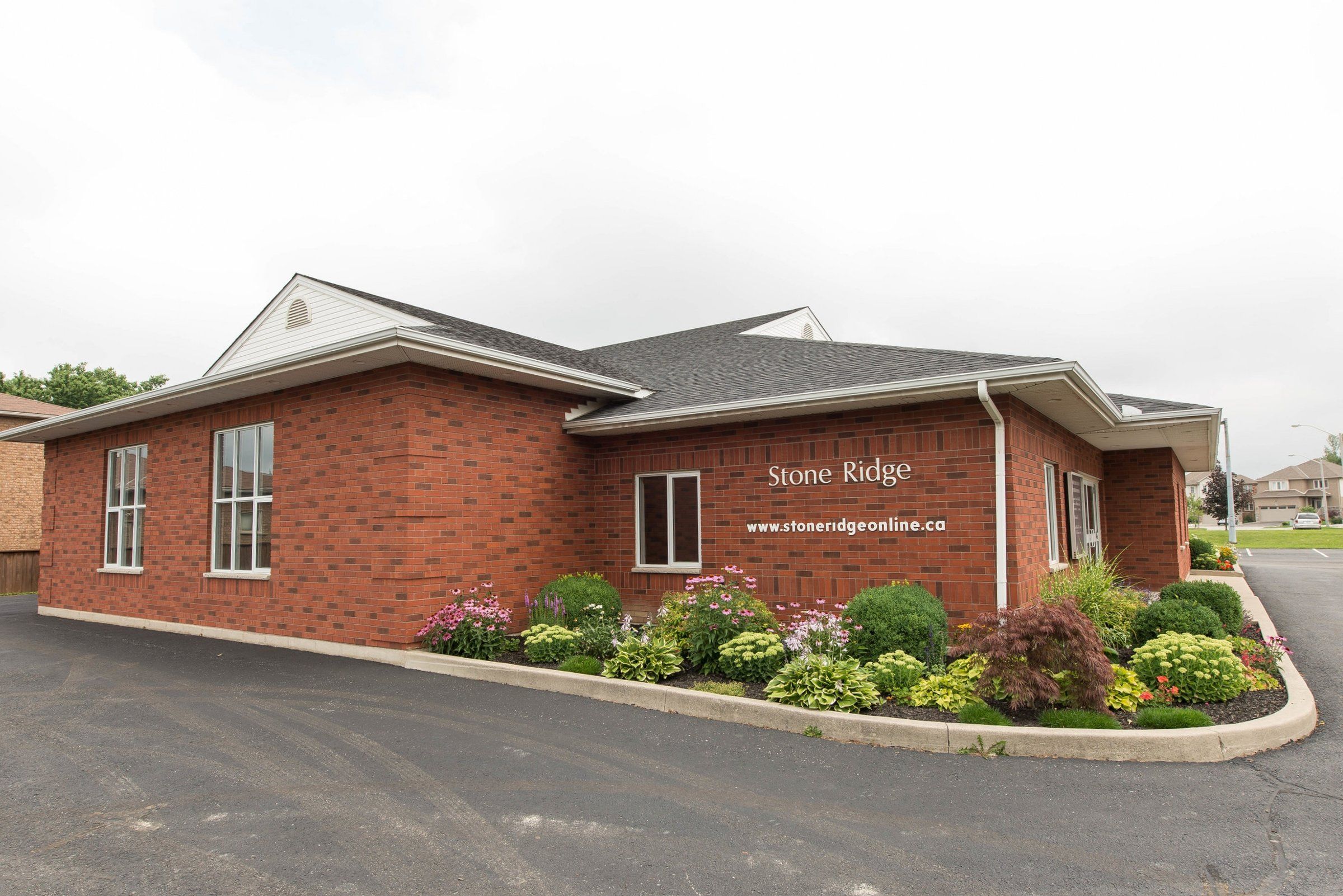Stone Ridge Bible Chapel, a single-storey red-brick building with a dark shingled roof and white trim, its wall lettered Stone Ridge and the church's web address above a flower bed beside the parking lot