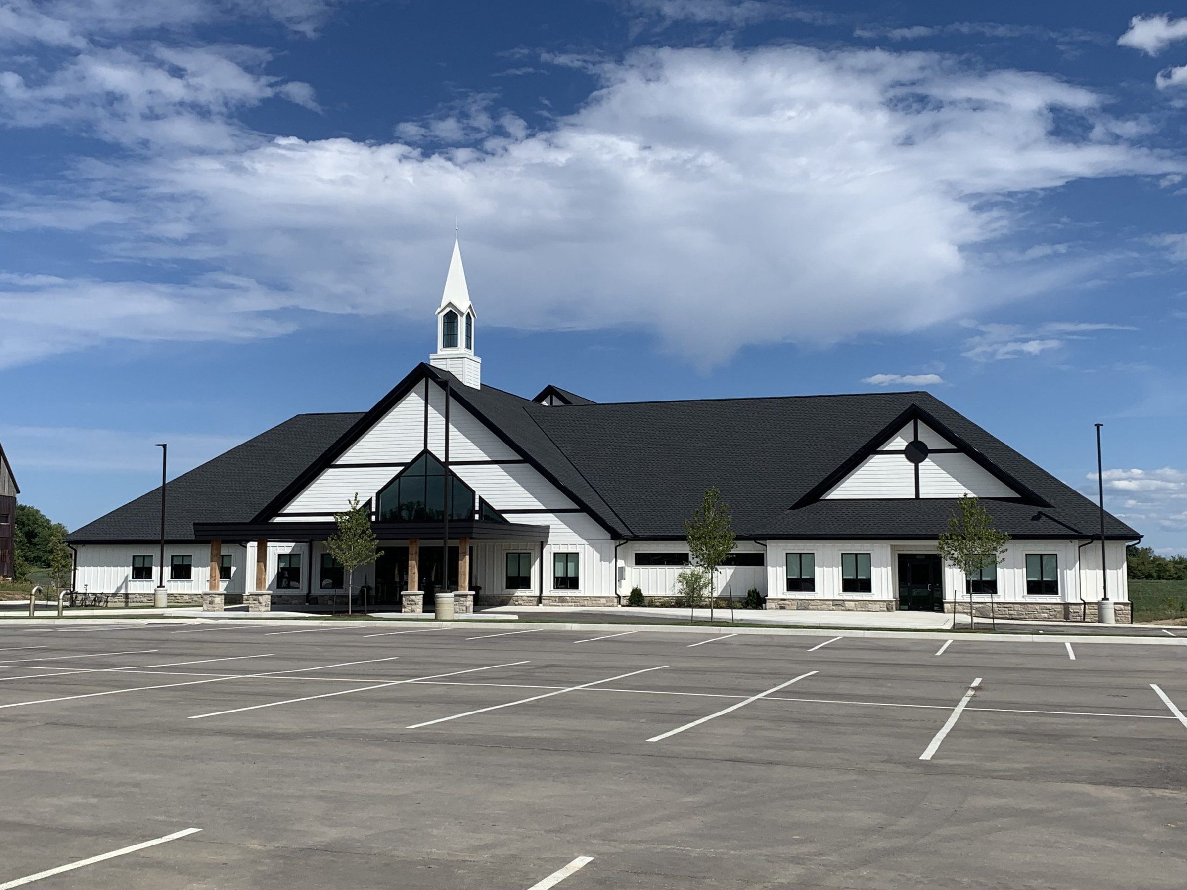 Trinity Canadian Reformed Church, a new white church with dark roofs, a tall glazed entrance under a steep gable and a slender white steeple, seen across its empty parking lot under a summer sky