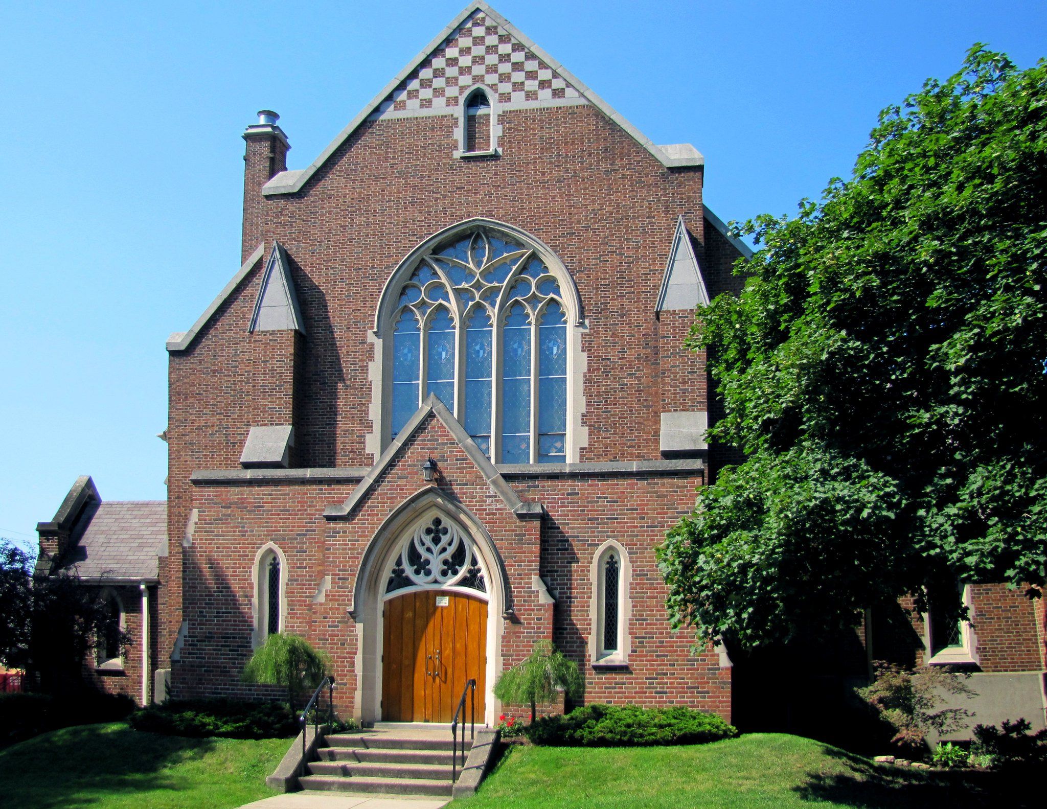 Westdale United Church, a red-brick church with a large pointed window filled with pale tracery above an arched wooden door, a chequered pattern high in its gable, and trees at its side